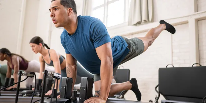 a man wearing a blue shirt lifts his leg to hold a pose on a Pilates Reformer machine