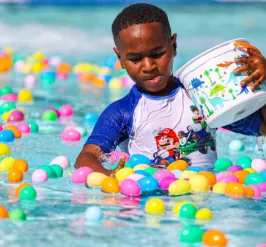 Boy in wet swimming clothes holding a basket in the pool surrounded by colorful plastic eggs