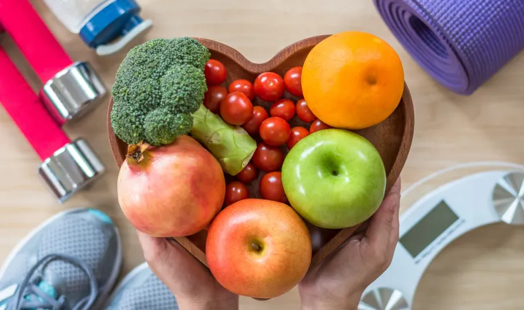 collection of fruits and vegetables in a heart-shaped bowl, held over pieces of fitness equipment